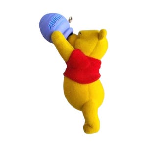 May include: A yellow bear figurine with a fuzzy texture wearing a red shirt. The bear is reaching upward with both arms to hold a light blue, rounded container that has a textured surface and a small silver metal loop attached to the top.