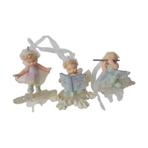 May include: Three small cherub ornaments with glittery accents and white ribbon hangers. One cherub stands, one sits reading a blue book labeled 'Little Ones' with '2005' printed on the cover, and one sits playing a silver flute-like instrument.