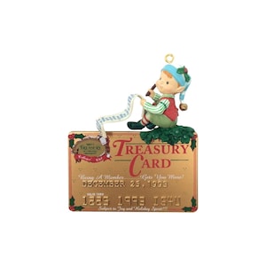 Könnte beinhalten: Ein Weihnachtsschmuck mit einem kleinen Elf mit blauer Mütze und roter Weste, der auf einer goldenen, rechteckigen Treasury Card sitzt. Die Karte zeigt den Text Treasury Card, December 25, 1993, und die Nummer 1225 1993 1540, verziert mit grünem Stechpalmenlaub.