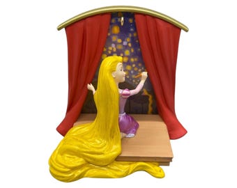Peut inclure: Une figurine décorative représentant un personnage aux longs cheveux jaunes vêtu d'une robe violette, assis sur une plateforme en bois entre des rideaux rouges. Le personnage tend la main vers un fond sombre rempli de petites lumières jaunes.