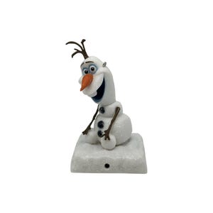 May include: A white, glittery snowman figurine with a carrot-shaped orange nose, brown twig-like arms and hair, and three black buttons on its torso. The snowman is seated on a square, glittery white base with a small circular hole in the front.