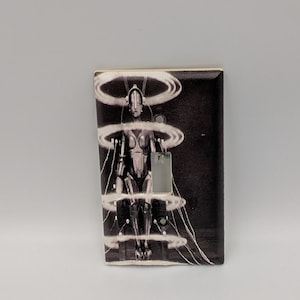 May include: A rectangular light switch cover plate featuring a black and white graphic print of a robotic figure surrounded by four glowing horizontal rings. The design includes thin white lines extending from the rings, with a central toggle switch cutout.