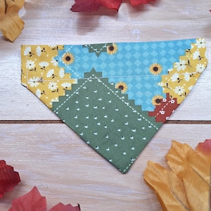 May include: A triangular pet bandana featuring a patchwork design with sections of yellow fabric with white daisies, light blue fabric with a small diamond pattern and sunflowers, and olive green fabric with a pattern of tiny white hearts.