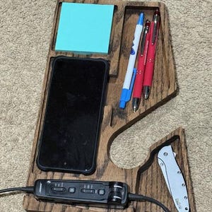 Könnte beinhalten: Schreibtisch-Organizer aus Holz mit Fächern für Telefon, Stifte, Notizblock und kleines Messer. Der Organizer ist aus dunklem Holz mit natürlicher Maserung gefertigt. Ein schwarzes Telefon und ein blauer Notizblock sind sichtbar.