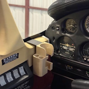 May include: Beige 3D-printed airplane accessory, likely a phone or cup holder, mounted near the dashboard. The dashboard features gauges, switches, and the text "WARNING". The interior is a mix of beige, black, and red.