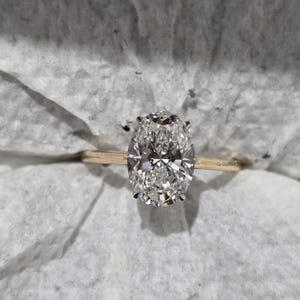 May include: A gold ring featuring a large, oval-cut, clear faceted gemstone held by four prongs. The band is thin and polished gold. The text "Grantha Design" is engraved on the side of the band near the setting of the oval gemstone.
