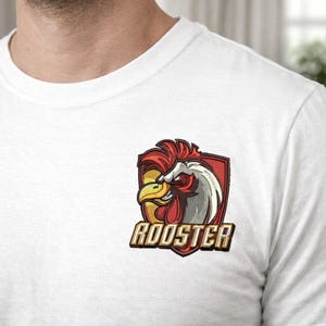 Pode incluir: Camiseta branca com um emblema de galo. O emblema tem a forma de um escudo com um desenho de galo vermelho, amarelo e branco e a palavra "ROOSTER" em letras douradas. A camiseta é de gola redonda.