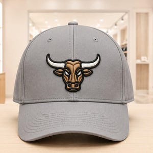May include: A light grey baseball cap featuring a centred, embroidered graphic of a bull head. The bull is rendered in shades of tan and brown with prominent white horns, outlined in black thread. The cap has a structured crown and a curved brim.