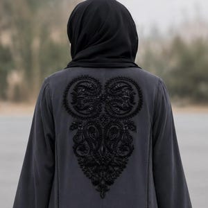 Peut inclure: Une veste gris foncé à manches longues avec un motif brodé noir complexe dans le dos. La broderie se compose de motifs cachemire et floraux détaillés avec des perles. Un couvre-chef noir uni est porté par-dessus la veste.