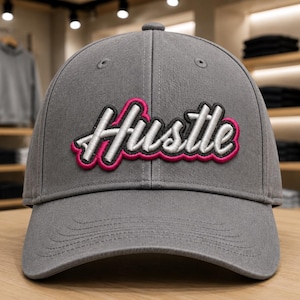 May include: A grey baseball cap featuring the word Hustle embroidered in white script with a thick black outline and a vibrant pink border. The cap has a structured crown, a curved brim, and matching grey stitching across the front panels.