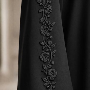 May include: A black garment sleeve featuring a vertical strip of tonal black floral embroidery. The embroidery depicts a winding vine with detailed leaves, small buds, and blooming flowers, all rendered in matching black thread on the fabric.