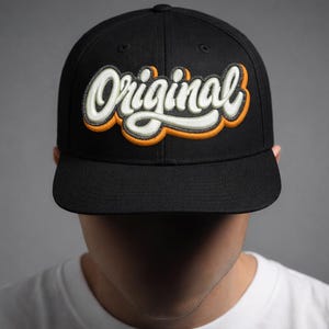 May include: Black baseball cap featuring the word "Original" embroidered in white with orange and grey detailing. The cap has a curved peak and a structured design. The person is wearing a white t-shirt.