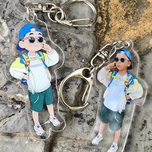 May include: Two clear acrylic keychains featuring a cartoon illustration of a child wearing sunglasses, a blue cap, and a backpack. The child is dressed in a white shirt, green shorts, and white sneakers. The keychains have silver-colored clasps.