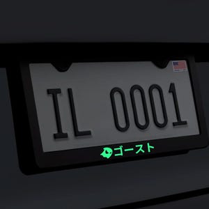 Puede incluir: Placa de matrícula con las letras 'IL' y el número '0001'. El marco tiene texto japonés y un gráfico de fantasma verde brillante. Se ve una pequeña bandera americana.