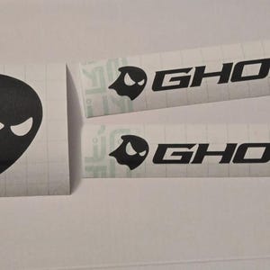 Pode incluir: Um conjunto de decalques em preto e branco com um logotipo fantasmagórico e a palavra "GHOST". Os decalques incluem um logotipo quadrado e dois decalques retangulares com o logotipo e o texto.
