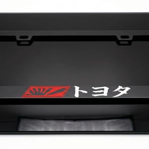 May include: A black rectangular license plate frame featuring a red graphic of a rising sun on the left side and the white Japanese text Toyota on the right side. The frame has four mounting holes at the top and bottom for vehicle attachment.