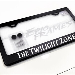 Peut inclure: Cadre de plaque d'immatriculation noir avec le texte "THE TWILIGHT ZONE" en blanc. Le cadre comprend le matériel de montage et est sur une surface blanche. Le cadre est rectangulaire et a des coins arrondis.