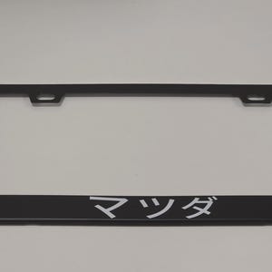 Puede incluir: Marco de matrícula negro con la palabra japonesa "Mazda" en letras blancas. El marco tiene forma rectangular con orificios de montaje en la parte superior.