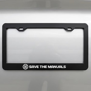 May include: Black license plate frame with the text "SAVE THE MANUALS" in white. The frame has a rectangular shape with rounded corners and mounting holes. The frame is mounted on a silver surface.
