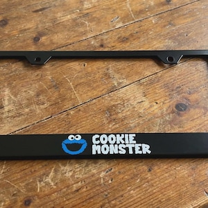 May include: Black license plate frame featuring the Cookie Monster character with blue and white details. The text "COOKIE MONSTER" is displayed in white. The frame is rectangular and has mounting holes.
