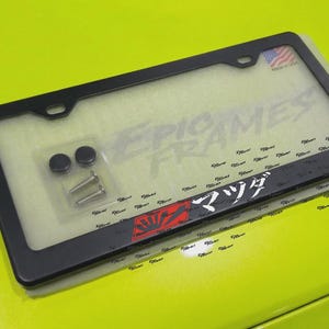 以下が含まれることがあります： 「EPIC FRAMES」の文字と日本語の文字が入った黒いナンバープレートフレーム。取り付け金具付き。小さなアメリカ国旗のステッカーが付いています。フレームは明るい黄色の表面にあります。