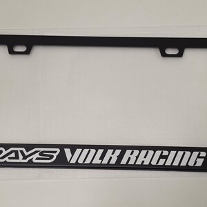 Puede incluir: Marco de matrícula negro con el texto blanco "RAYS VOLK RACING". El marco rectangular tiene dos orificios de montaje en la parte superior. El marco está hecho de un material negro mate.
