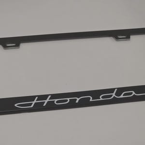 May include: Black car license plate frame with the white word "Honda" in a cursive font. The frame is rectangular with rounded corners and has mounting holes at the top.