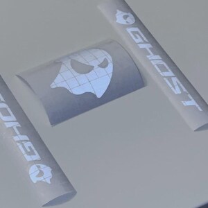 May include: White vinyl stickers with the word "GHOST" in a bold font, along with a ghost icon. The stickers are in a roll and flat sheet. The ghost icon has a spooky face. The stickers are on a white surface.