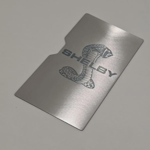 以下が含まれることがあります： 前面に「SHELBY」の文字とコブラのロゴが刻まれた、銀色の金属製カードホルダー。 カードホルダーは、片側に切り欠きのあるユニークな形状をしています。