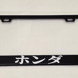 May include: Black license plate frame with the Japanese characters for "Honda" in white lettering. The frame is rectangular with mounting holes at the top and is displayed on a white background.