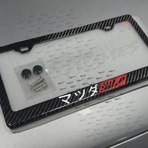 Op de afbeelding: Een zwart kentekenplaatraam van koolstofvezel met het Japanse woord "Mazda" in het wit en een rood-wit logo. Inclusief montagemateriaal.