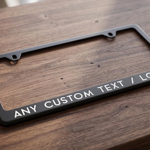 Puede incluir: Un marco rectangular negro para matrícula con esquinas redondeadas y dos orificios de montaje en la parte superior. El borde inferior del marco presenta el texto impreso en blanco ANY CUSTOM TEXT / LOGO en una fuente sans-serif.
