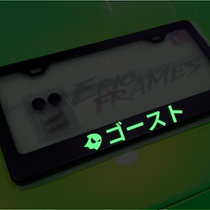 May include: Black license plate frame with the Japanese word "GHOST" and a ghost icon, both glowing green. The frame also has the text "EPIC FRAMES". Includes mounting hardware.