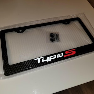 May include: Black license plate frame with a carbon fiber design. The frame has the text "Type S" in white and red. Includes mounting hardware.