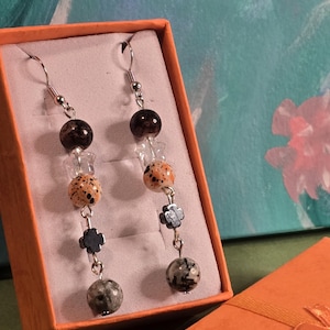 May include: A pair of dangle earrings displayed in an orange gift box. Each earring features a dark brown bead, clear crystal beads, speckled orange beads, a silver-toned clover charm, and a gray and black marbled bead. The earrings are on a white background.