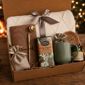 Könnte beinhalten: Eine Geschenkbox gefüllt mit einer cremefarbenen Decke, einem braunen Lederjournal, einem goldenen Stift, einer Tasse und einem Schokoriegel. Die Box ist mit einem grauen Band gebunden und enthält einen kleinen Jutesack und einen goldenen Löffel.