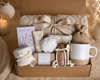 May include: A gift box filled with self-care items. Includes a white mug with a tan base, a beige knitted blanket, a bar of soap, a small bag, a jar of bath salts, a white flower, and a tube of lotion. The box is tied with a beige ribbon.