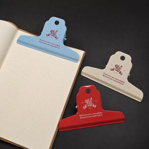May include: Three metal binder clips in light blue, beige, and red, each featuring a circular hole at the top and the printed text "Discover your serendipity Record your inspirations" with a small graphic logo above the lettering.