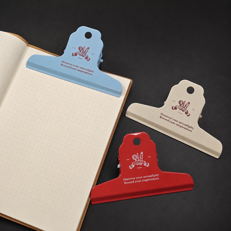 May include: Three metal binder clips in light blue, beige, and red, each featuring a circular hole at the top and the printed text "Discover your serendipity Record your inspirations" with a small graphic logo above the lettering.