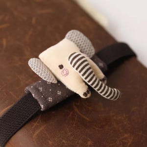 May include: A fabric elephant bookmark featuring a cream-colored head, gray textured ears, and a brown and white striped trunk. The elephant is attached to a dark brown elastic band with a small gray fabric sleeve featuring a white dotted pattern.