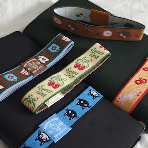 May include: Four elastic bands for notebooks featuring various patterns: a brown band with mugs, a beige band with cherries and the text "Sweet Time", a blue band with black cat faces, and a rust-colored band with white daisies and brown accents.
