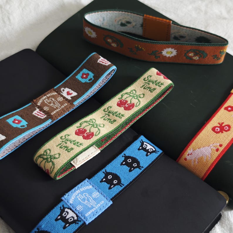 May include: Four elastic bands for notebooks featuring various patterns: a brown band with mugs, a beige band with cherries and the text "Sweet Time", a blue band with black cat faces, and a rust-colored band with white daisies and brown accents.