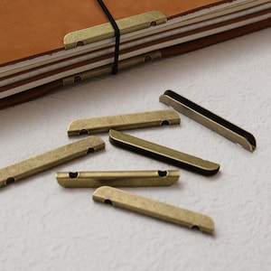 May include: A collection of antique brass-colored metal book spine protectors with a U-shaped profile and small circular cutouts at each end, scattered alongside a brown leather-covered notebook secured with a black elastic band.