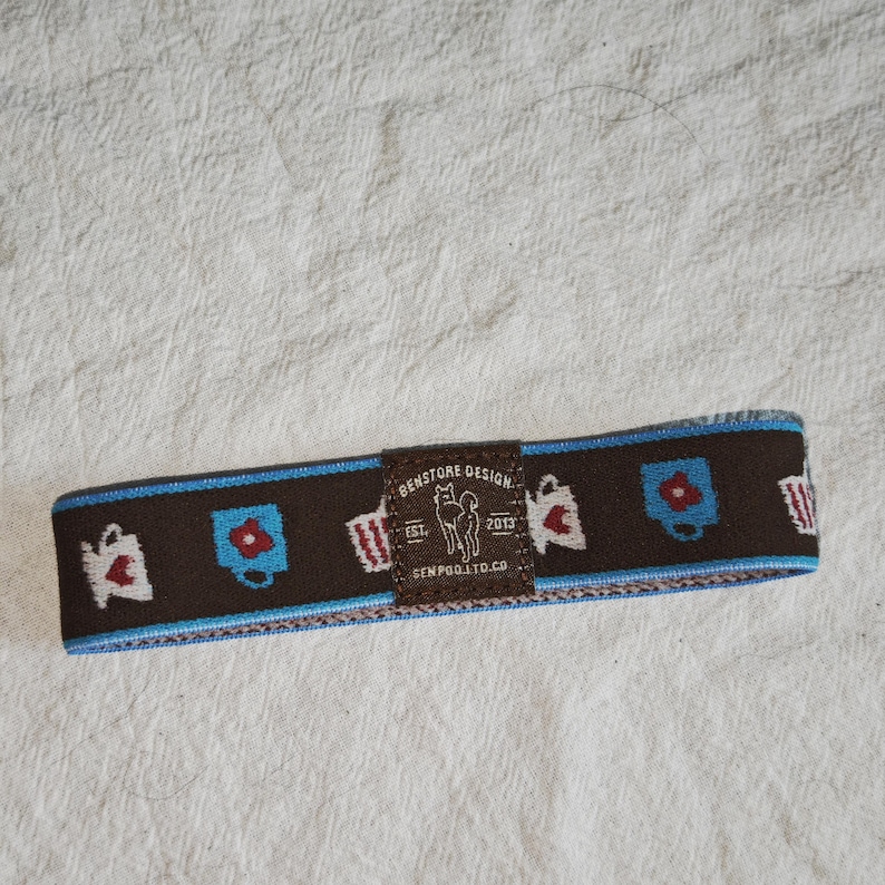 May include: A brown elastic headband featuring a repeating pattern of small white and blue mugs with red accents. A central brown rectangular patch displays the text BENSTORE DESIGN EST. 2013 SENPOO.LTD.CO with a small dog illustration.