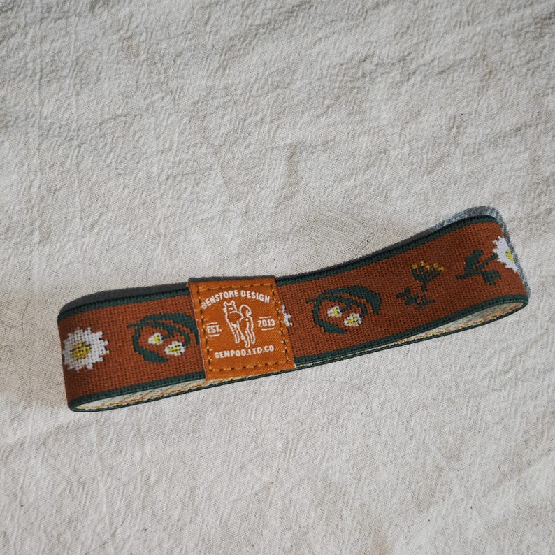 May include: A brown elastic headband featuring a repeating pattern of white daisies and dark green leaves. A central tan leather patch displays the text "BENSTORE DESIGN EST. 2013 SENPOO.LTD.CO" along with a line drawing of a dog silhouette.