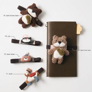 May include: A collection of elastic bands featuring plush animal figures including a dark brown bear, a cat, a bear head, a duck, and a light brown bear attached to a dark brown leather notebook. Labels identify items 10, 11, 12, 13, and 14.