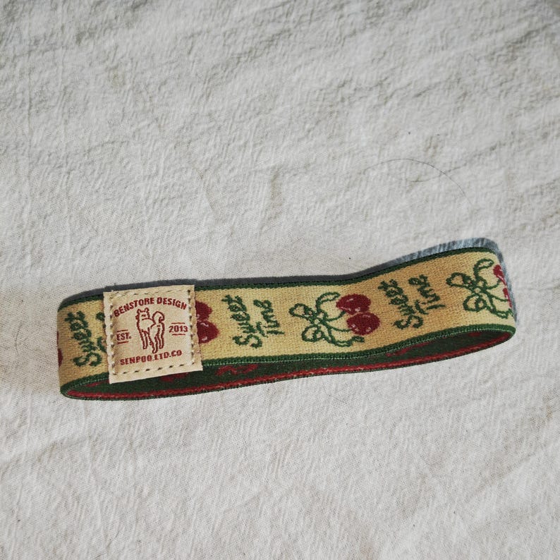 May include: A beige elastic headband featuring a repeating pattern of red cherries and green leaves with the text "SWEET TIME" in dark green. A rectangular fabric label is attached, displaying a dog illustration and the text "BENSTORE DESIGN EST. 2013 SENPOO.LTD.CO".