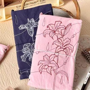 May include: Two fabric notebook covers, one in dark navy blue and one in light pink, each featuring white or dark red embroidered floral designs of lilies and raindrops, secured with a thin white elastic band wrapped around the center.