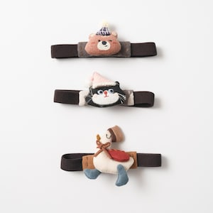 May include: Three elastic hair bands featuring fabric appliques. The top band has a brown bear wearing a blue and white striped hat. The middle band features a black cat with a pink hat. The bottom band displays a white goose with a brown hat.