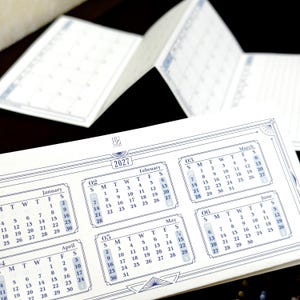 May include: A white accordion-fold paper planner for the year 2027 with blue ink printing. The planner features a grid layout for the months of January through June, with the text HUGO PLANNER visible at the top center of the page.